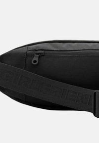 Borsetă din țesătură neagră, cu un compartiment principal prevăzut cu fermoar și o curea reglabilă cu numele mărcii embossat.