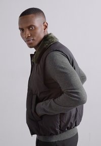 Črn podložen brezrokavnik z zelenim umetnim krznom, z bočnimi žepi in prilegajočim krojem, oblečen preko sivega dolgosečnega majice.