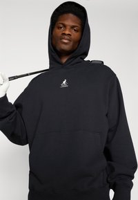 Sweatshirt preto com capuz feito de uma mistura de algodão. Possui um bolso tipo canguru, punhos canelados e um pequeno logótipo no peito. Textura suave.