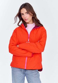 Kobieta z długimi brązowymi włosami, ubrana w pomarańczową pikowaną kurtkę na różowym swetrze i jasnoniebieskich dżinsach, z rękoma skrzyżowanymi na piersi, na neutralnym tle.