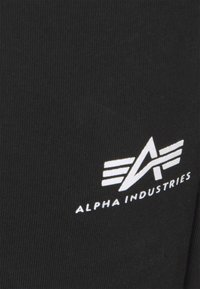 Sort stof med en ribbet tekstur, der præsenterer et hvidt logo af "Alpha Industries" samt et emblem med vinger og et stiliseret "A."