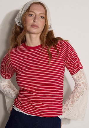 Donna con lunghi capelli rossi che indossa una maglia a righe rosse e bianche sopra maniche bianche di pizzo e un fazzoletto bianco di pizzo in testa.