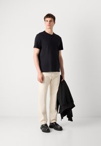 Black short-sleeve T-shirt, complemented by cream trousers. The individual holds a black, zippered jacket, wearing black shoes with a textured surface.