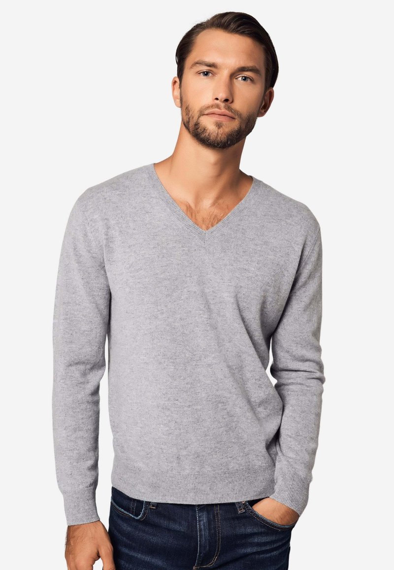 Mann mit Bart, der einen hellgrauen V-Ausschnitt-Pullover und dunkle Jeans trägt, steht mit einer Hand in der Tasche vor einem einfarbigen Hintergrund.