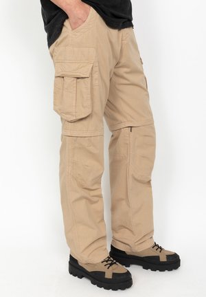 Mężczyzna w beżowych, odpinanych spodniach typu cargo i jasnobrązowych butach z czarną podeszwą, stojący z jedną ręką w kieszeni.