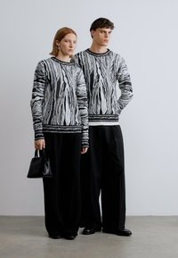 Pulls en maille à motifs noirs et blancs avec manches longues, associés à un pantalon large noir. L'une tient un petit sac à main noir.