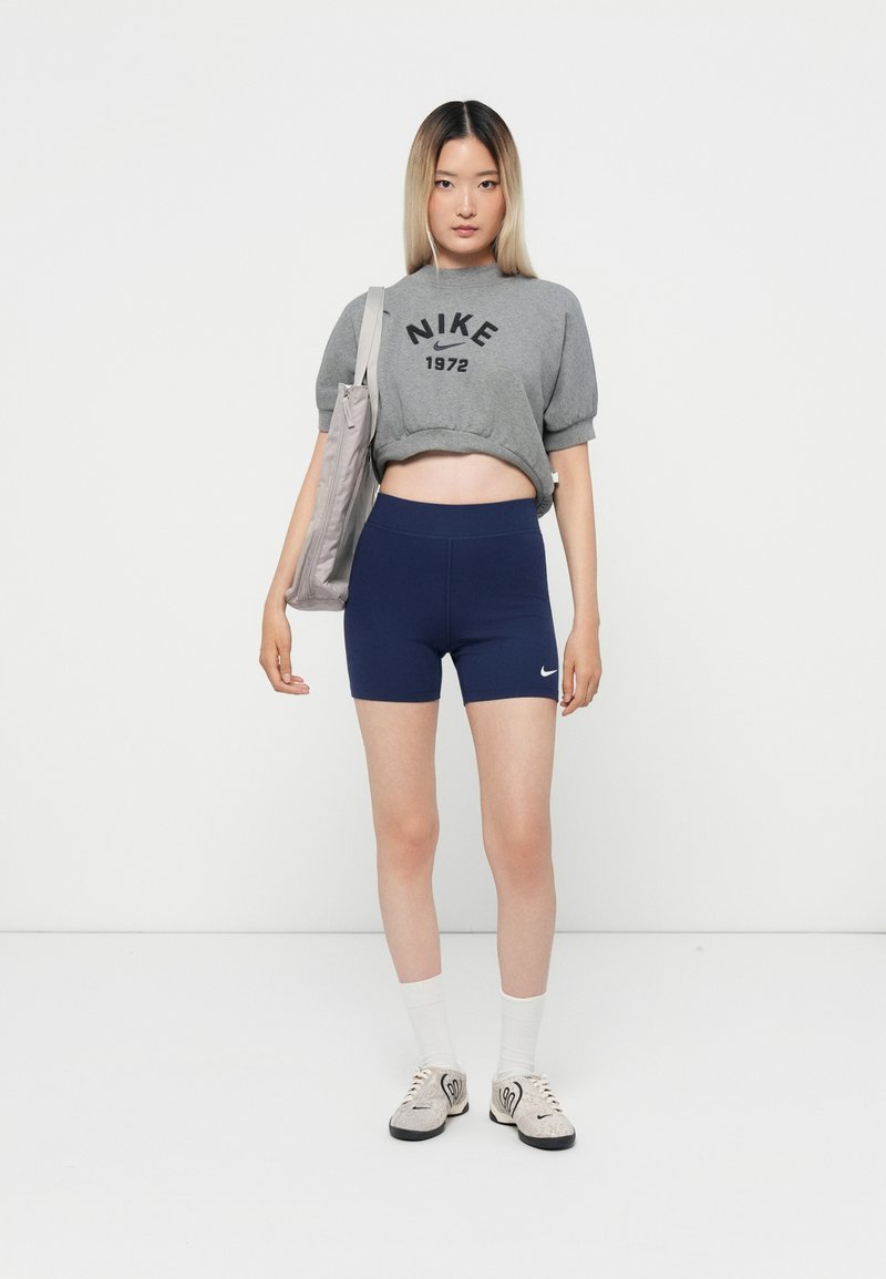 Sweatshirt gris court avec l'impression noire "Nike 1972", associé à un short de vélo marine, des chaussettes blanches et des baskets grises. Tenant un tote léger.