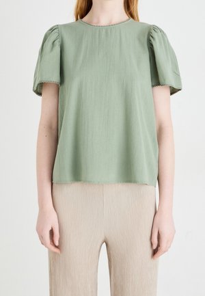 Femme portant un chemisier court à manches bouffantes vert clair et un pantalon beige texturé sur un fond clair uni.