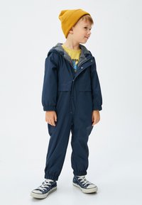 Marinevandtæt jumpsuit med elastiske manchetter, grå foret hætte og trykknapper, båret med en gul hue og blå sneakers.