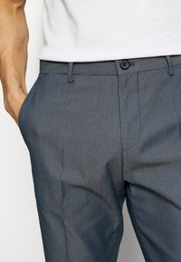 Kalhoty v námořnické modři s geometrickým bílým vzorem, se zapínáním na knoflík a kapsami. Látka se zdá být hladká s přiléhavým střihem.