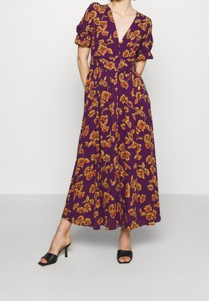 Kobieta ubrana w fioletową sukienkę midi w żółto-czerwony kwiatowy wzór, z bufkami na rękawach, paskiem w talii i czarnymi sandałami na obcasie z odkrytymi palcami.