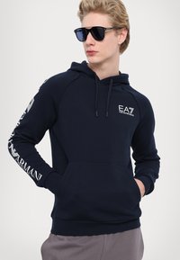 Sötétkék kapucnis pulóver elülső kenguruzsebbel, zsinórral és fehér EA7 logóval a mellkason és az ujján. Sima textúra, laza tervezés.