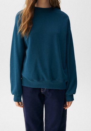 Kvinde iført teal langærmet sweater med ribkantede ærmer og mørkeblå jeans mod en ensfarvet lys baggrund.