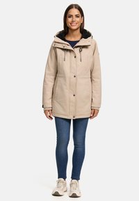 Parka imperméable beige avec une capuche amovible en fourrure noire, fermeture éclair à l'avant, boutons-pression et deux poches latérales. Portée sur un jean bleu.