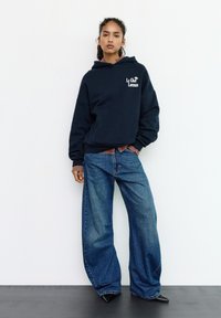 Felpa con cappuccio blu navy con tasca frontale e testo grafico, abbinata a jeans blu a gamba larga. Scarpe nere con punta a punta completano l'outfit.