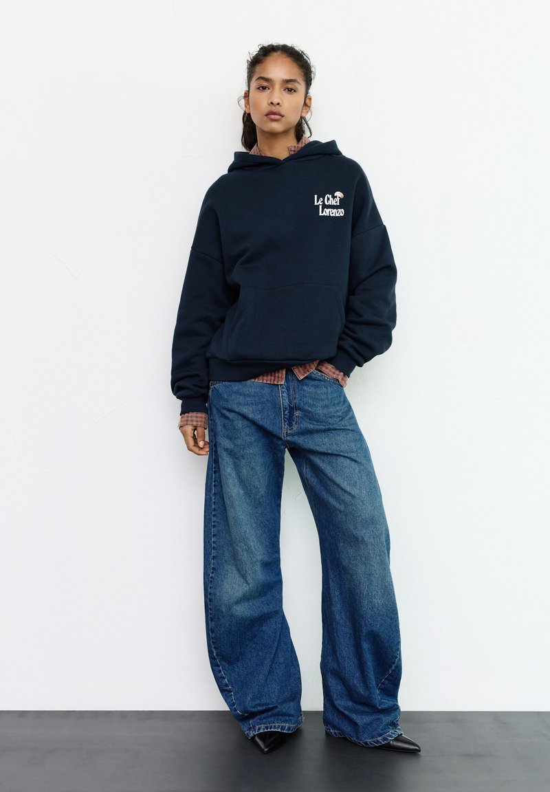 Felpa con cappuccio blu navy con tasca frontale e testo grafico, abbinata a jeans blu a gamba larga. Scarpe nere con punta a punta completano l'outfit.