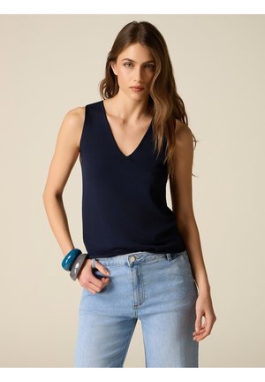 Femme aux longs cheveux bruns portant un haut bleu marine sans manches à col en V, un jean bleu clair et des bracelets épais bleus et gris au poignet gauche.