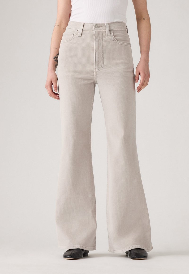 Personne portant un pantalon évasé taille haute beige clair, des chaussures noires et un débardeur blanc, debout devant un fond uni.