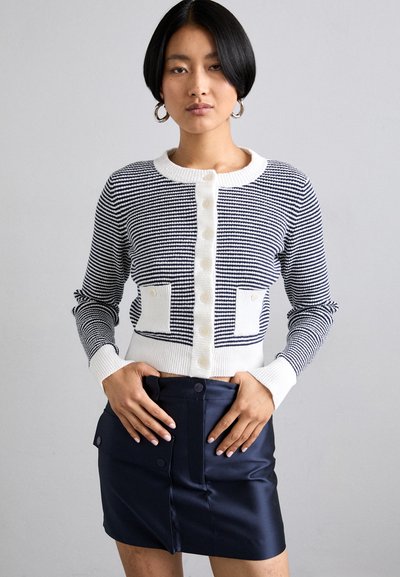 Marineblå og hvitstripete cardigan med knapper, ribbede mansjetter og to frontlommer. Kombinert med et høyt liv marineblå skjørt.