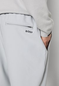 Pantalon gris clair avec une poche arrière zippée et un logo noir "BOSS", porté avec un haut à manches longues gris clair assorti.
