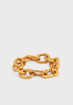 Pulsera de cadena gruesa dorada con grandes eslabones rectangulares y un cierre grabado con "MARC JACOBS" sobre un fondo claro.