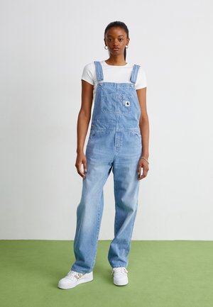 Salopette en denim bleu clair avec des bretelles ajustables, une poche avant et des jambes larges, associée à un t-shirt blanc et des baskets blanches.