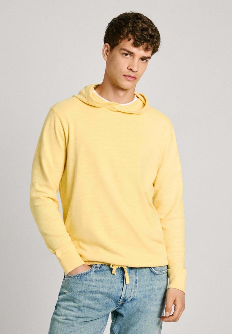 Sudadera amarilla con cordón, de manga larga, textura suave, combinada con jeans de denim azul claro. Diseño simple y casual.