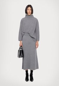 Grå strikket sweater med høj krave, parret med en matchende lang nederdel. Modellen holder en lille sort håndtaske. Sorte ankelstøvler fuldender looket.