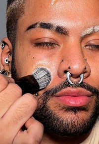 Person som applicerar smink med en borste på en ljus hudton. Framhävda ansiktspiercingar, inklusive septum, och kort mörkt hår. Nära fokus.