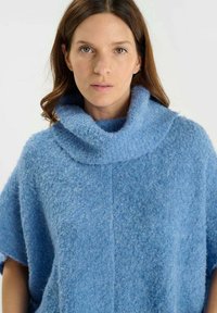 Donna con pelle chiara e capelli castani che indossa un maglione a collo alto blu strutturato, rivolta di fronte con espressione neutra su sfondo bianco.