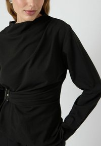 Haut noir à manches longues avec un col montant, une taille ceinturée et une texture lisse. Présente des épaules structurées et une coupe décontractée.