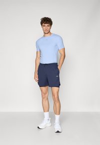 Hellblaues, tailliertes Sportshirt mit kurzen Ärmeln, kombiniert mit dunkelblauen Shorts mit Kordelzug und seitlichen Taschen. Weiße Sneakers.