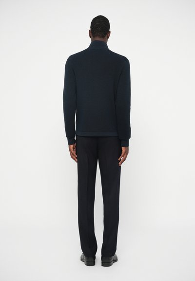 Pulover tricotat bleumarin cu guler înalt, asortat cu pantaloni închiși. Țesătura are o textură, oferind un contrast față de pantalonii netezi și bine croiți.