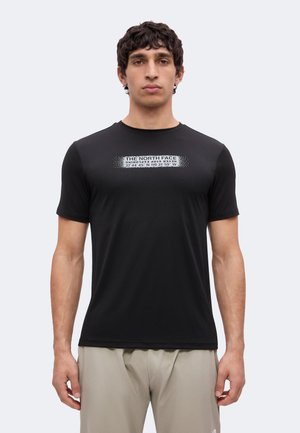 Mann trägt schwarzes The North Face T-Shirt mit Koordinaten und Text auf der Brust, beige Hose, steht vor einfachem hellem Hintergrund.