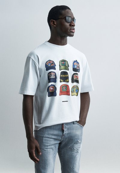 Άνδρας που φορά γυαλιά ηλίου, λευκό T-shirt με εννέα διαφορετικά χρωματιστά σχέδια καπέλων και ανοιχτό μπλε τζιν με σκισίματα, στέκεται μπροστά σε απλό φόντο.