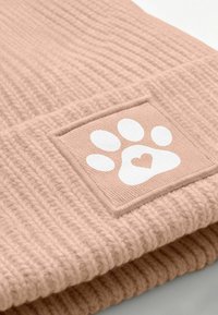 Pink knitted beanie features a textured pattern and a square patch with a white paw print and heart design. Soft material, snug fit.