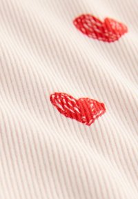 Puuvillakangas, jossa on pehmeä pinkki ribbitekstuuri, ja pinnalla on levitettyinä punaisia brodeerattuja sydämiä.