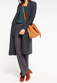 Cardigan long en maille gris foncé porté sur un haut turquoise, associé à un pantalon gris rayé verticalement et des bottines couleur rouille. Sac bandoulière marron.