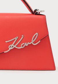 Red leather handbag with a triangular shape, featuring a silver metallic "Karl" logo accent and contrasting black stitching.