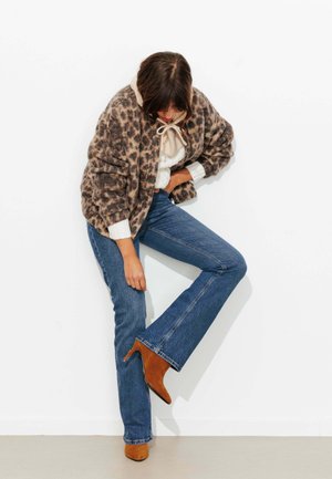 Femme portant un manteau à imprimé léopard, un pull blanc avec col à nœud, un jean bleu et des bottes à talons marron, levant une jambe contre un mur blanc.