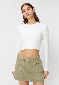 Biały, prążkowany crop top z długim rękawem w połączeniu z jasnooliwkową spódnicą dżinsową, która ma dwie przednie kieszenie i zapięcie na guziki.
