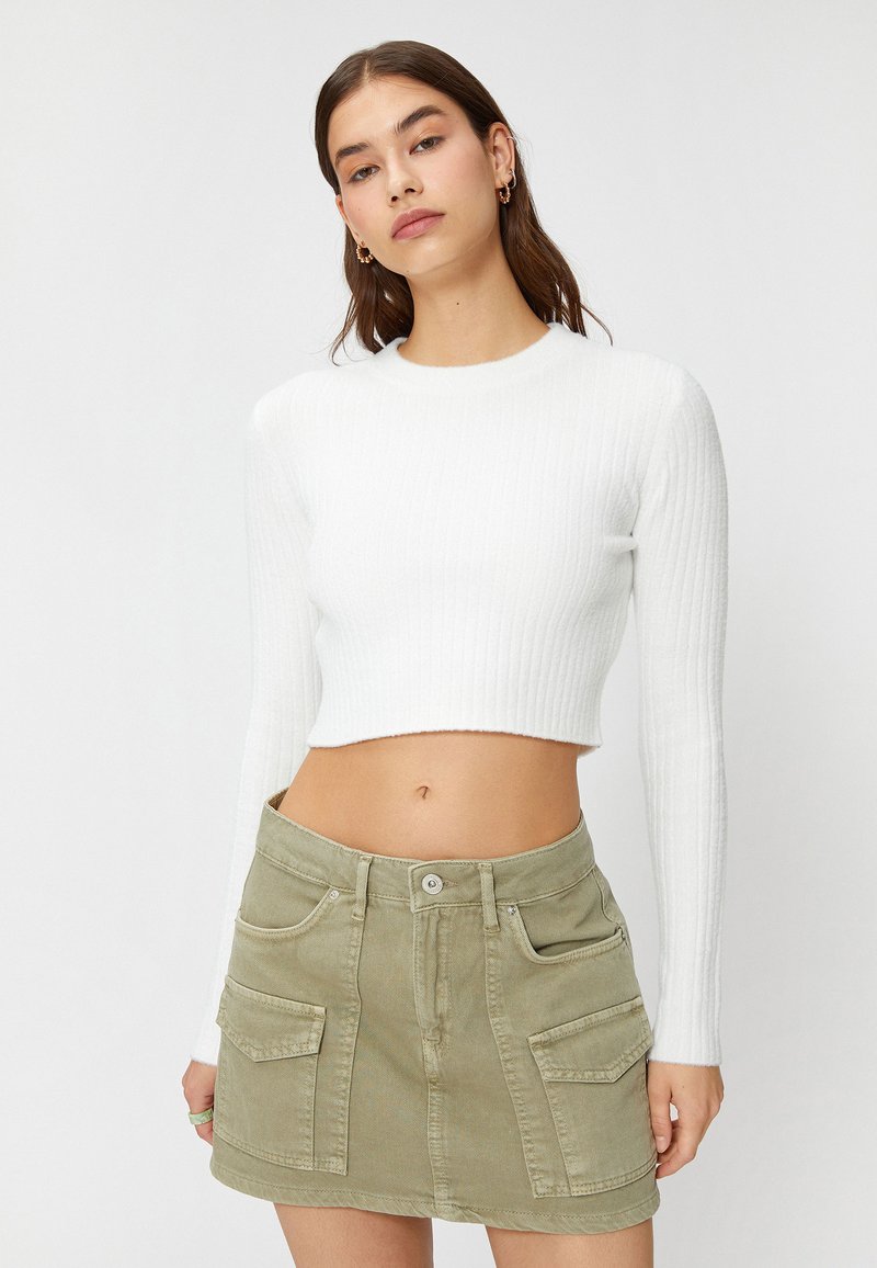 Biały, prążkowany crop top z długim rękawem w połączeniu z jasnooliwkową spódnicą dżinsową, która ma dwie przednie kieszenie i zapięcie na guziki.