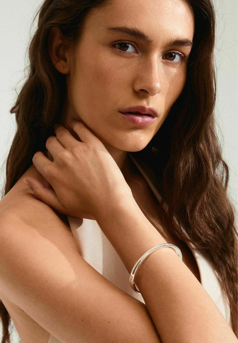 Bracciale in argento sul polso di un modello, texture liscia con una sottile lucentezza, poggiato su una pelle abbronzata, che mette in evidenza un design minimalista.
