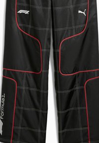Pantaloni de curse negri cu accente roșii, având cusături în carouri albe și logo-uri pentru Formula 1 și Puma. Materialul pare ușor și durabil.