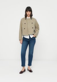 Kuttet beige dobbelspent jakke med mørke knapper. Kombinert med blå jeans og svarte flate sko. Modellen står med hendene i lommene.