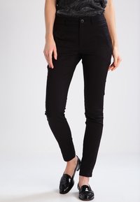 Pantalons noirs ajustés avec une texture lisse, poches latérales et fermeture par bouton. Associés à des mocassins noirs brillants.
