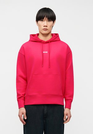 Persoon staat frontaal en draagt een felroze hoodie met voorzak en witte "MSGM"-logo, gecombineerd met donkere broek.
