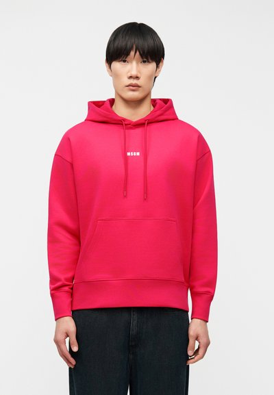 Personne debout, face à l'avant, portant un sweat à capuche rose vif avec une poche avant et le logo blanc "MSGM", associé à un pantalon foncé.