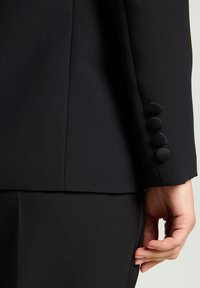 Schwarzer, maßgeschneiderter Blazer aus glattem Stoff mit geradem Schnitt, fünf dekorativen Knöpfen am Ärmel und einem eleganten, minimalistischen Design.