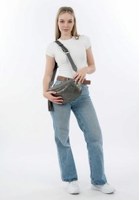 Borsa a tracolla in pelle grigia con chiusura a zip e dettaglio con frange, abbinata a jeans blu chiaro e una camicia bianca aderente.